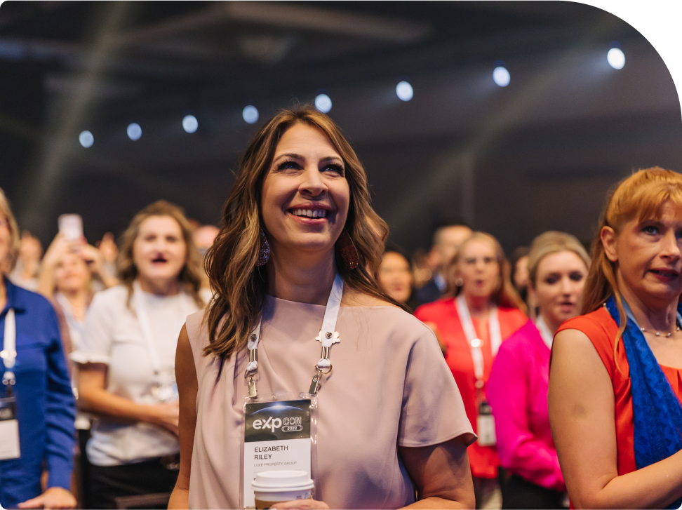 A woman smiling and standing in a crowd at a conference, wearing an eXp lanyard.